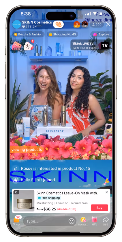 Two women smiling in a TikTok live stream promoting SKINN Cosmetics skincare products displayed on a table decorated with pink and red flowers.