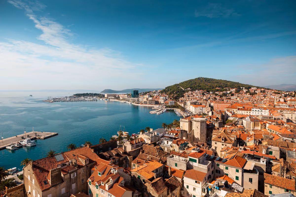Coastal city with red-roofed buildings by a harbor under a clear blue sky.