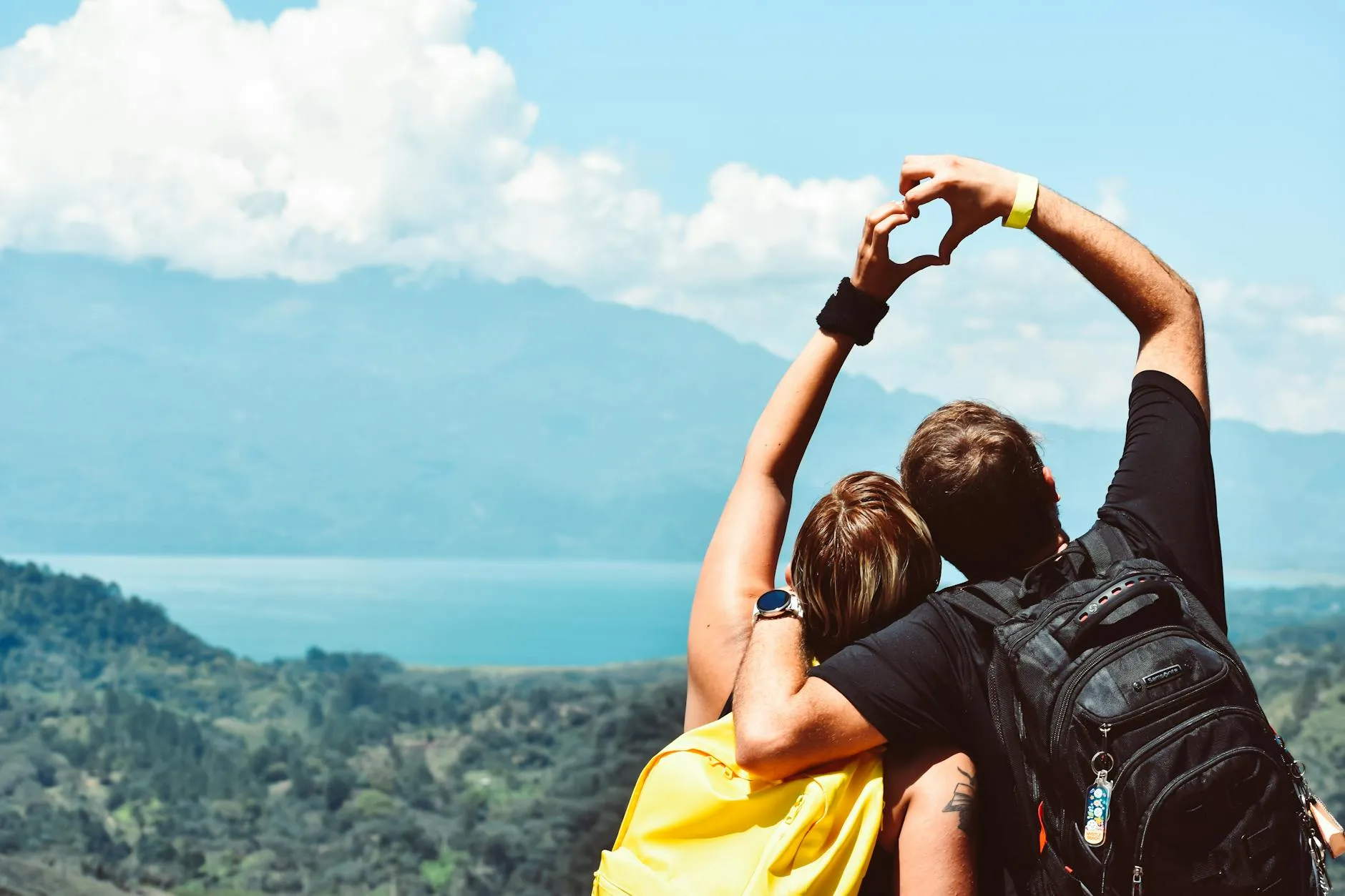 Couple with backpacks forming a heart shape with hands overlooking mountains and lake under a blue sky.