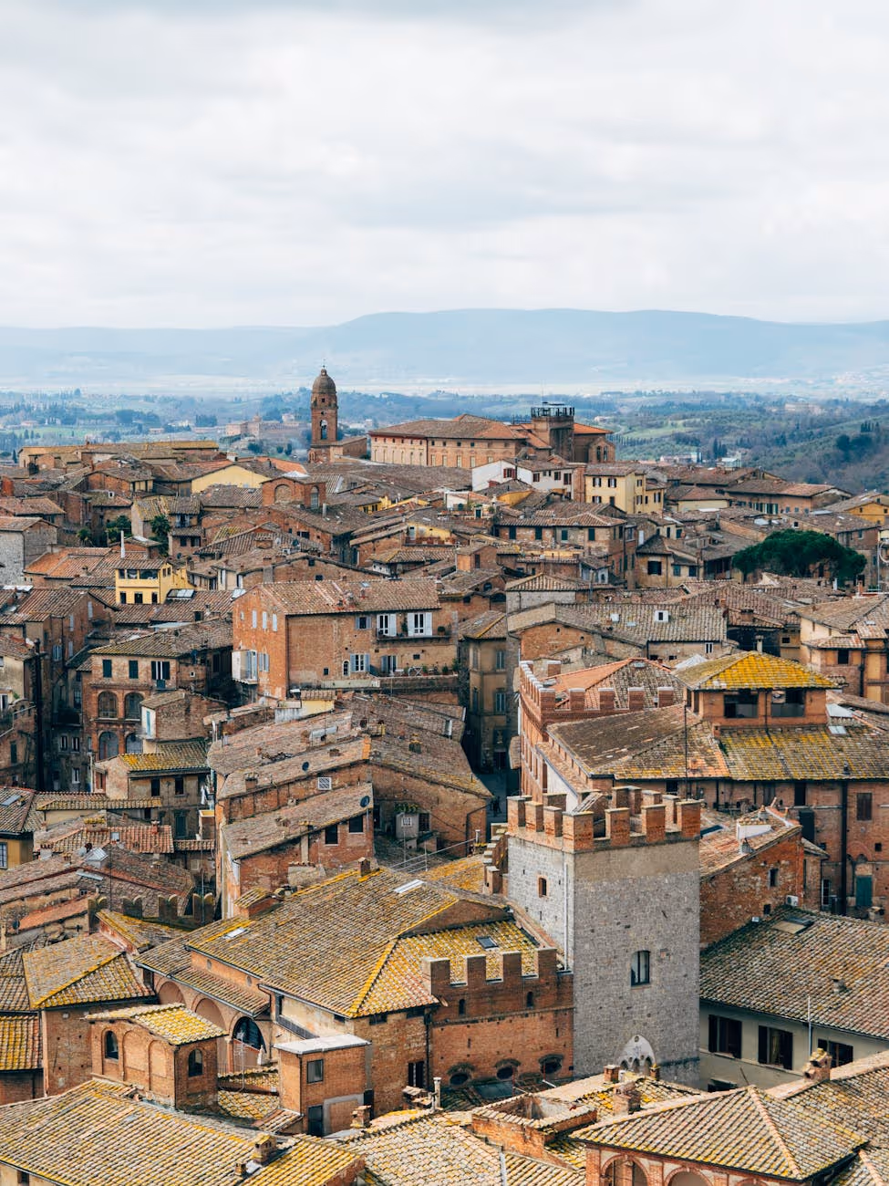 Aerial view of Tuscan town with terracotta rooftops, medieval buildings, and distant hills under cloudy sky.