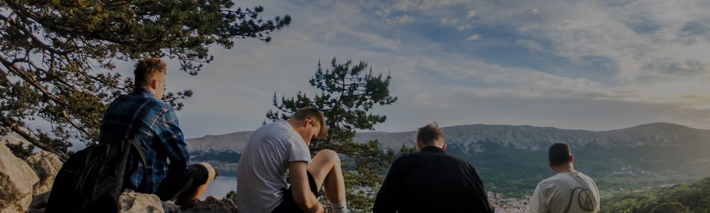 Four young men sitting on rocks overlooking a forested valley and mountains under a cloudy sky.