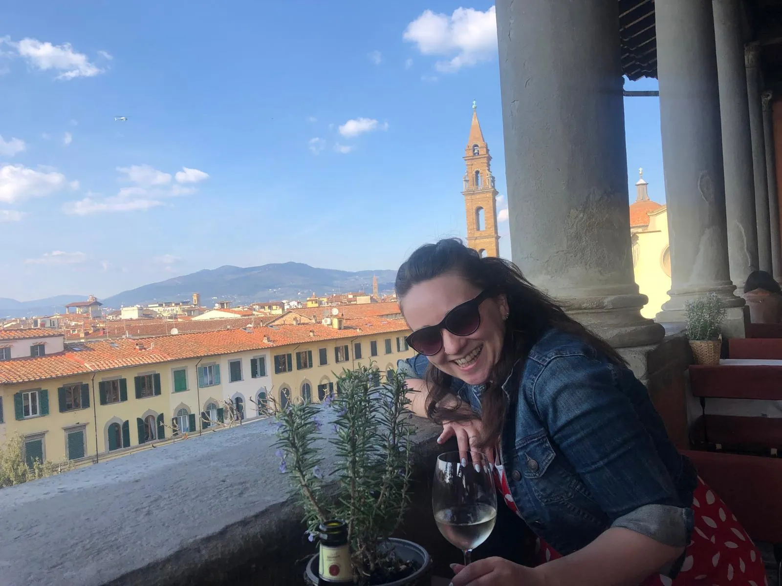 Smiling woman in sunglasses and denim jacket with a glass of white wine at an outdoor terrace.