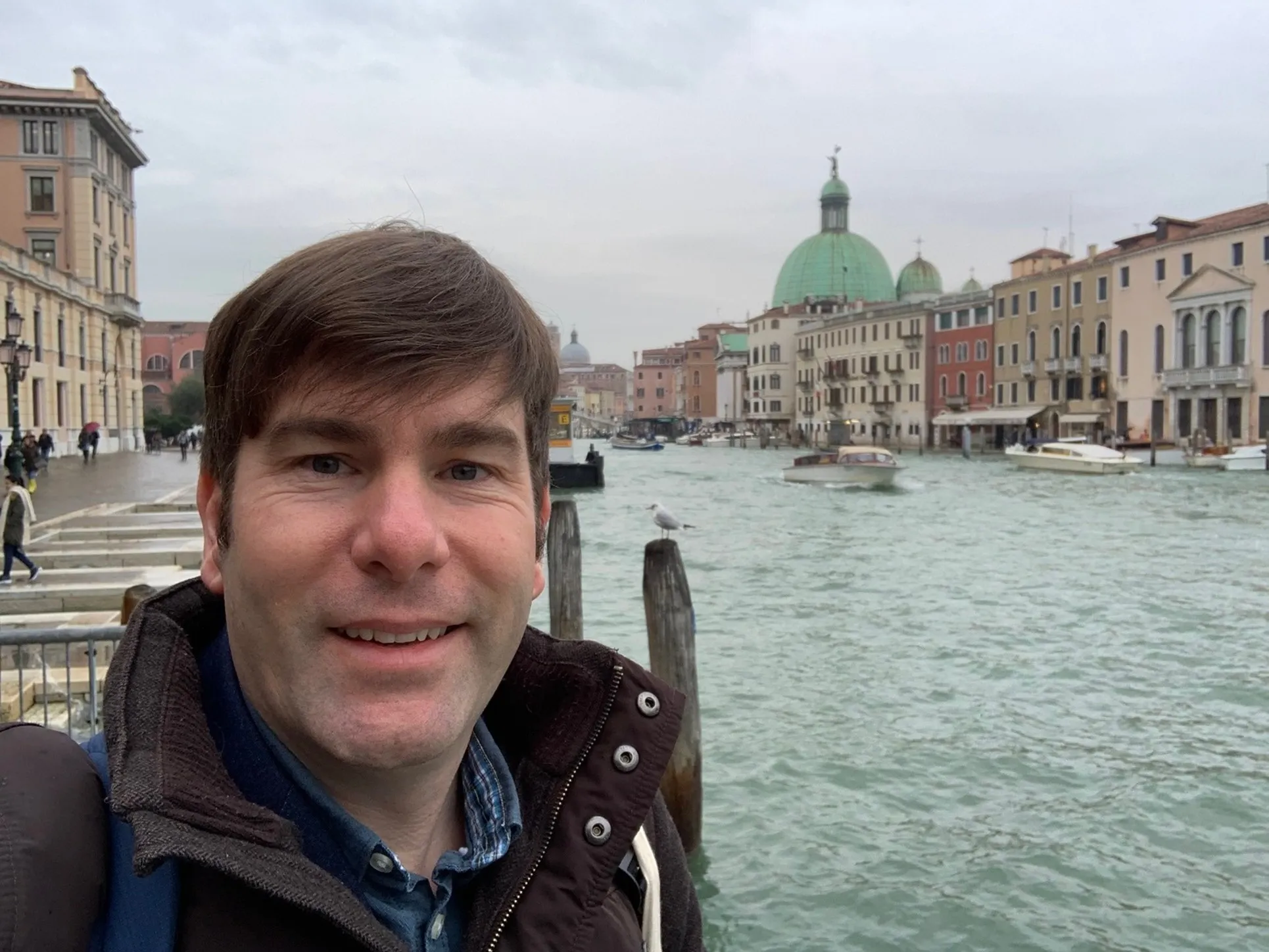 Man smiling with canal, boats, and historic buildings with domes in the background on a cloudy day.