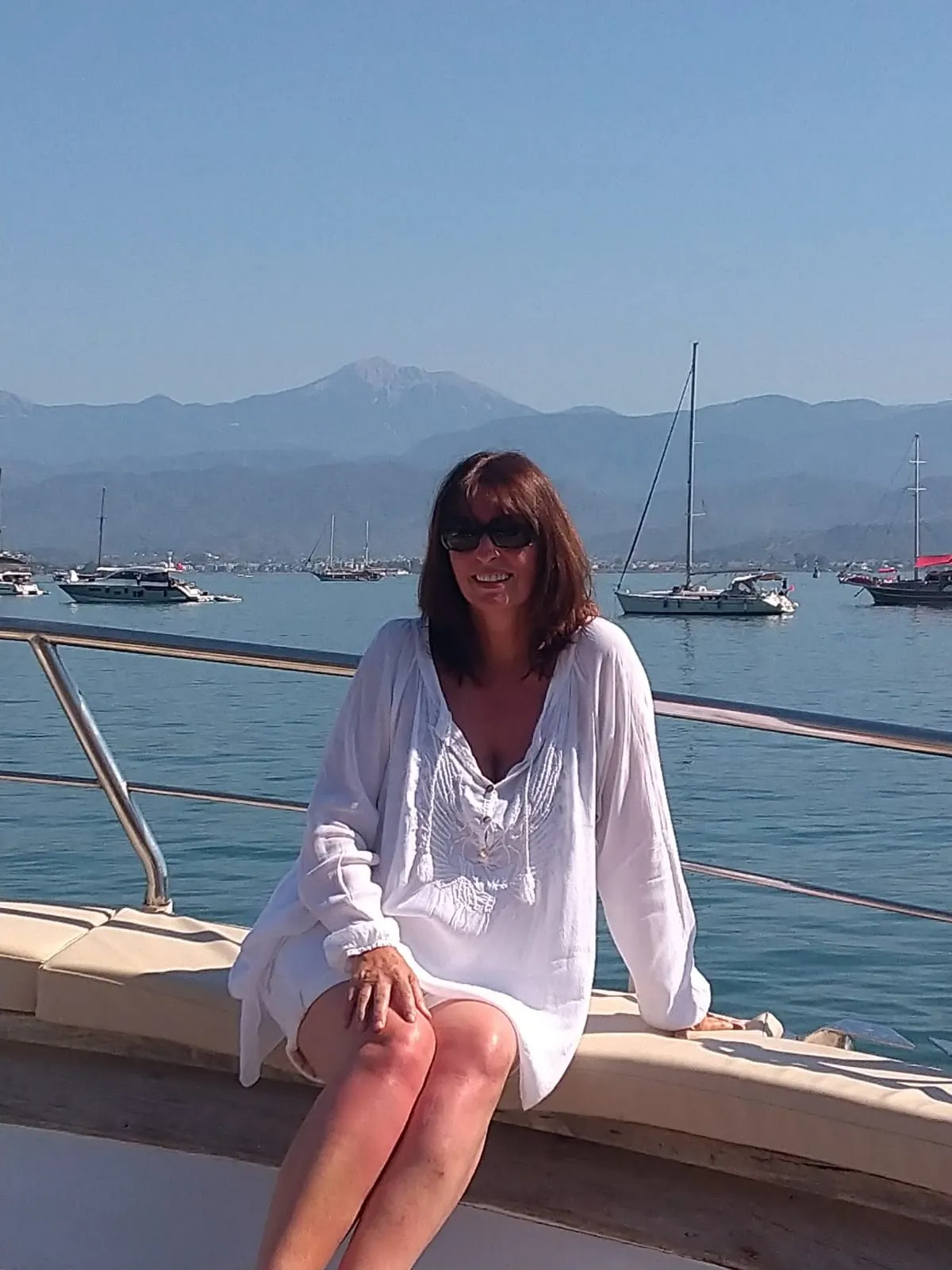 Woman in white dress and sunglasses sitting on boat railing with sea, boats, and mountains behind her.