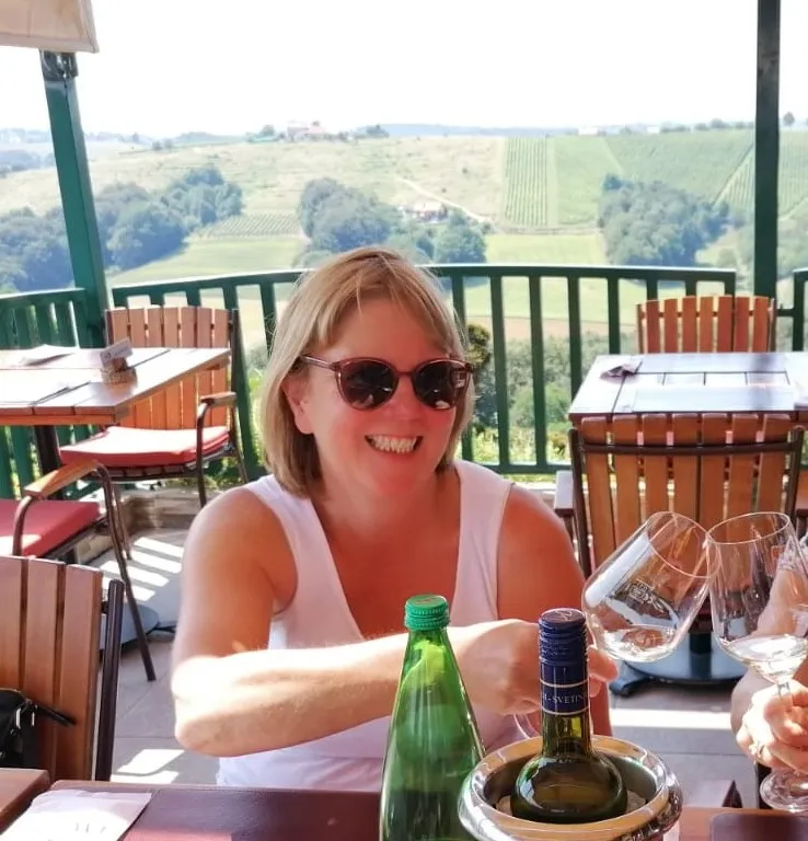 Smiling woman wearing sunglasses toasting wine glasses at an outdoor table overlooking vineyards.