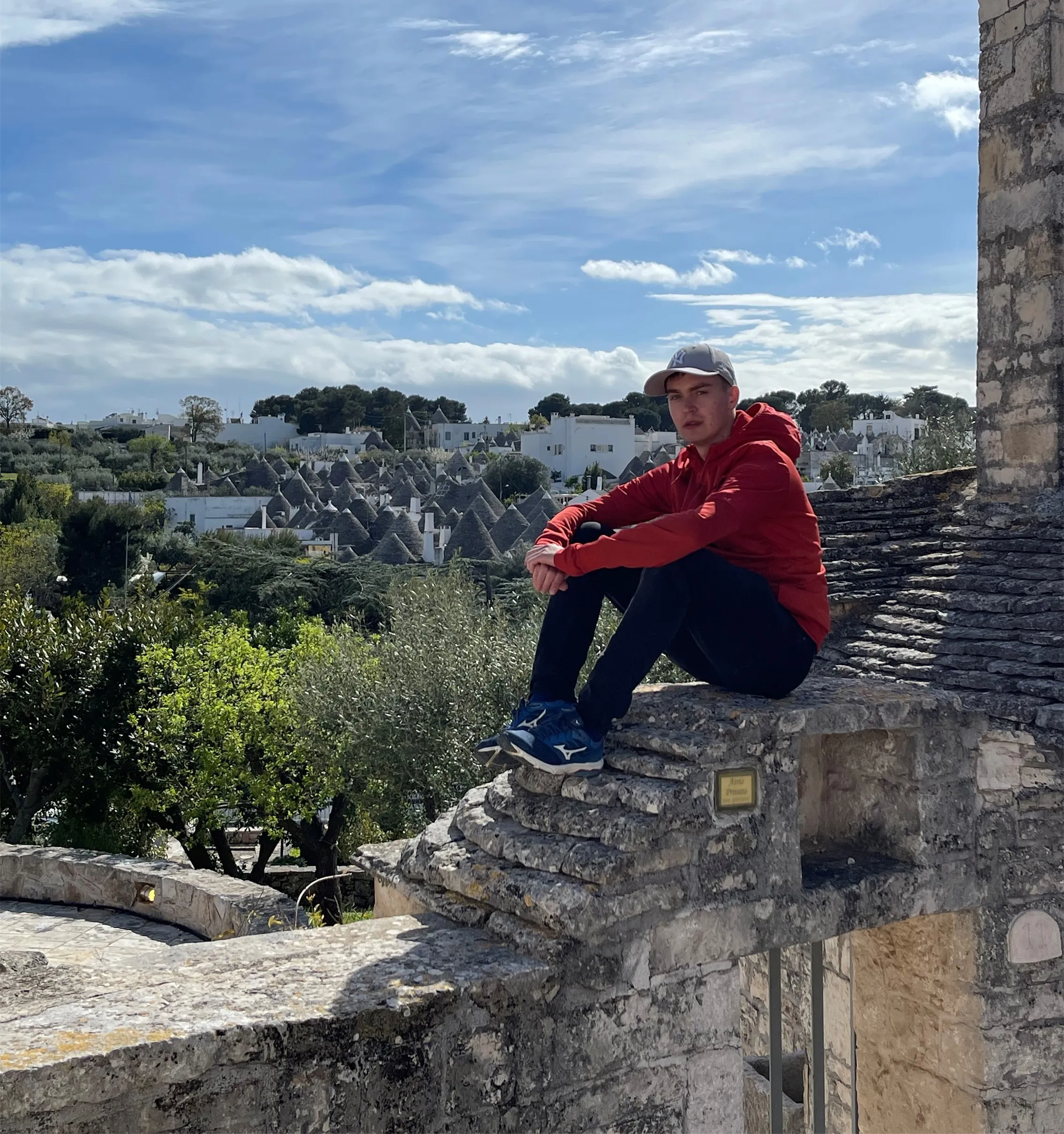 Young man in a red jacket and cap sitting on stone wall with historic cone-roof houses and blue sky behind.