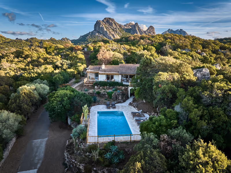 House with tiled roof surrounded by dense greenery and mountains, featuring a fenced rectangular pool.