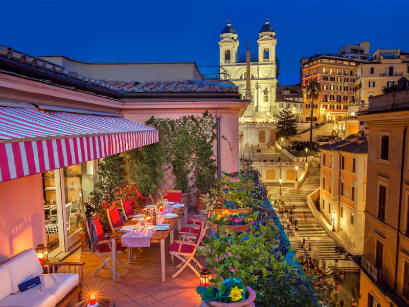 Evening view of a rooftop terrace set for dinner with red chairs and flowers overlooking historic city buildings.