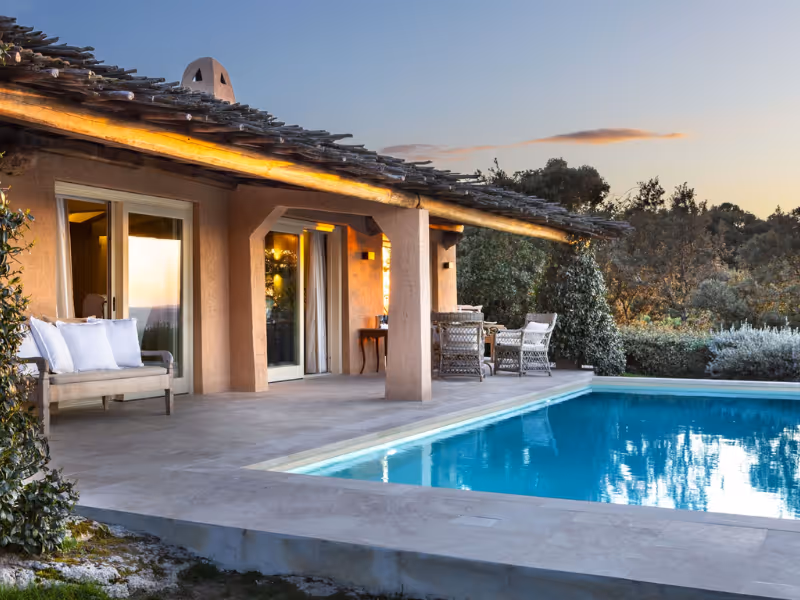Modern patio with seating next to a swimming pool at sunset with surrounding trees and bushes.