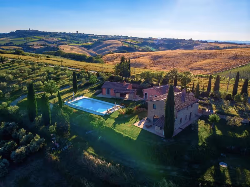 Stone villas with a swimming pool surrounded by tall cypress trees in a Tuscan hillside vineyard.
