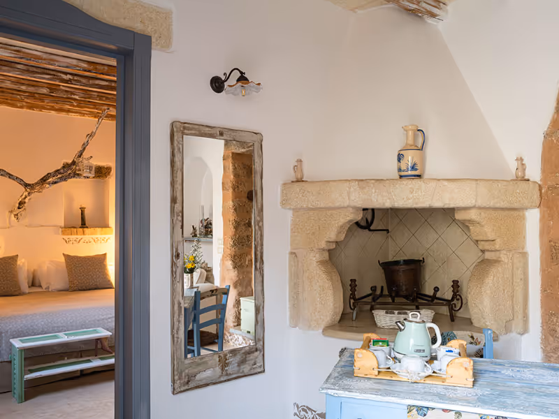 Cozy rustic room with stone fireplace, mirror, blue table with tea set, and a bed in adjoining room.
