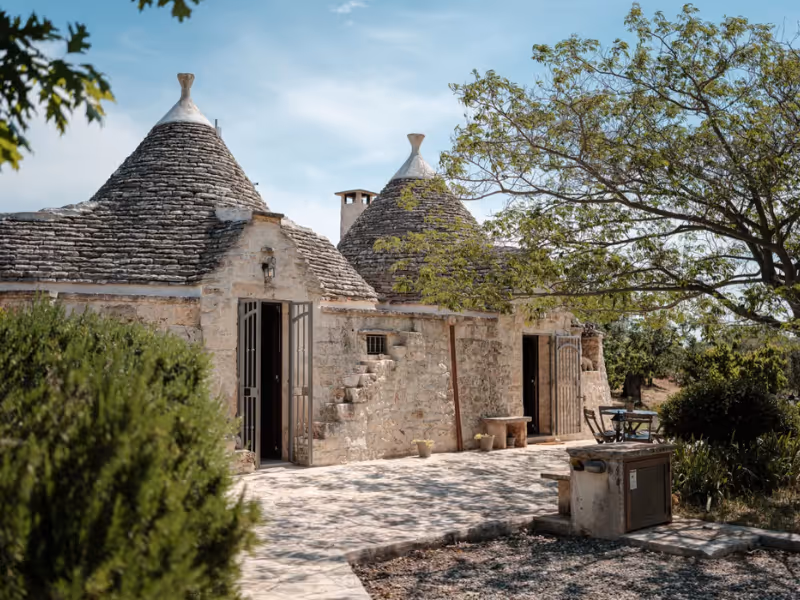Stone Trullo houses with conical roofs and open doors surrounded by trees and greenery under a blue sky.