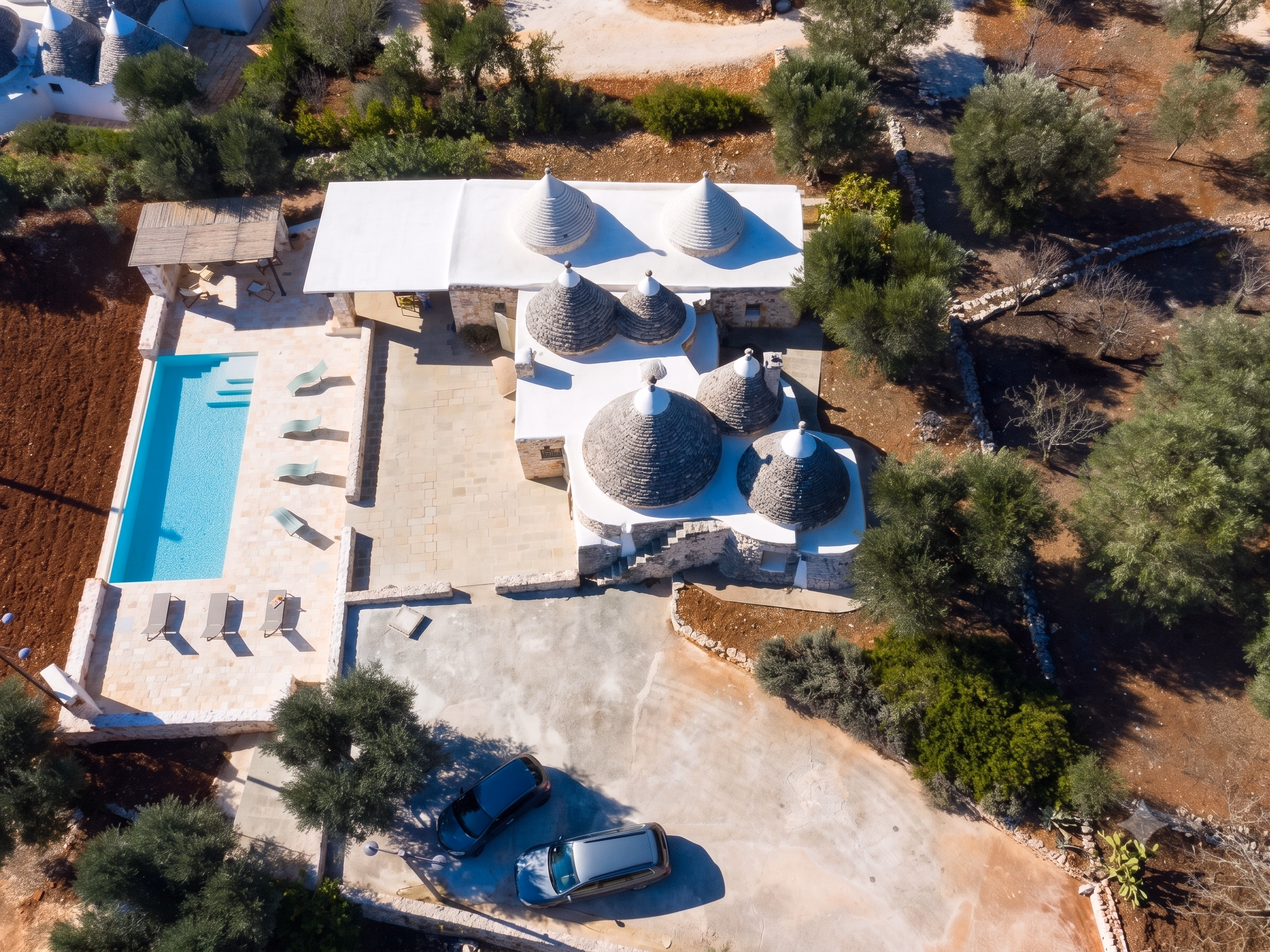Aerial view of a trullo-style stone house with conical roofs, a pool, trees, and two parked cars.