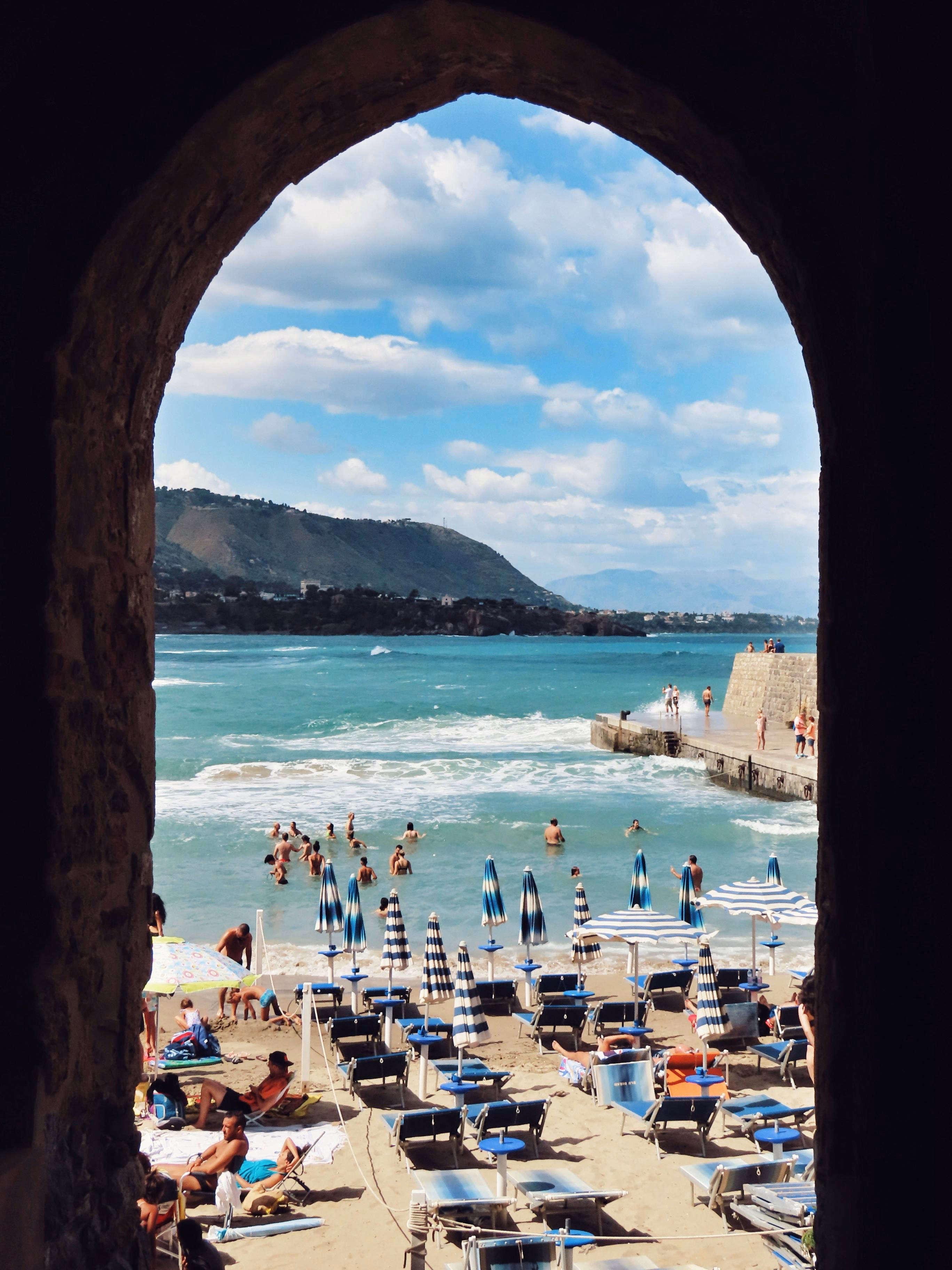 View of a crowded beach and sea with swimmers framed by a stone archway, mountains in background.