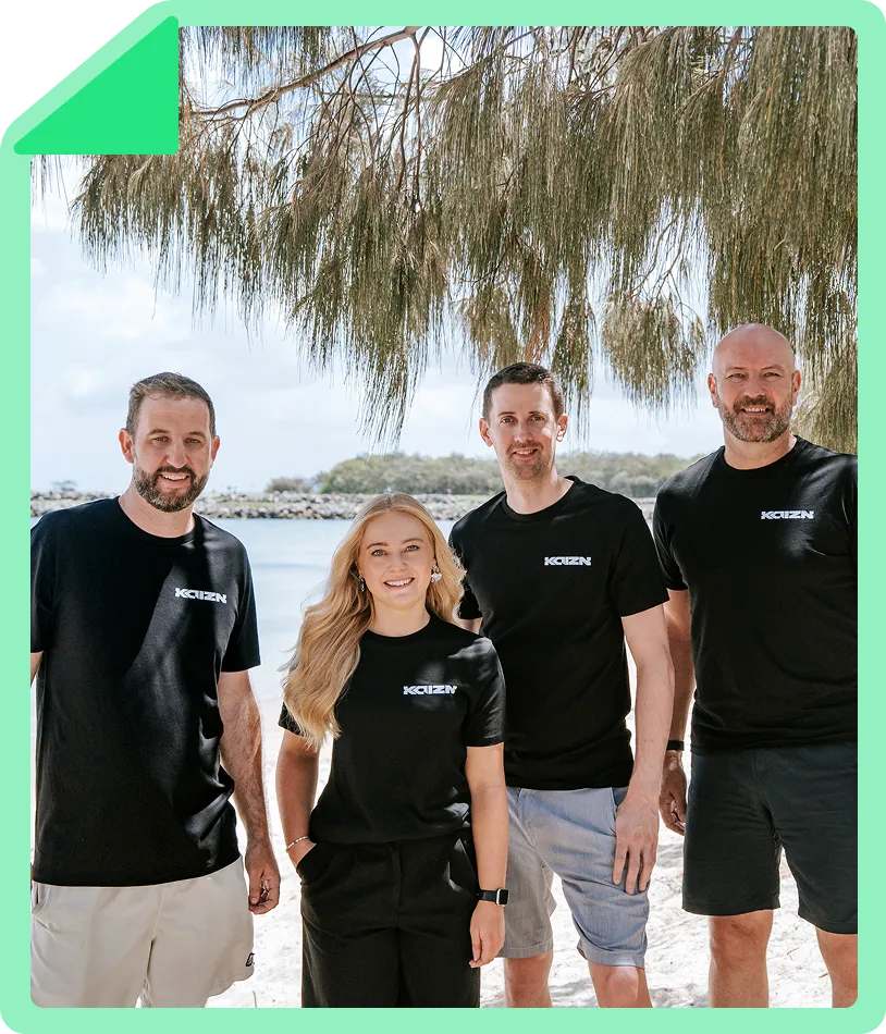 Four people wearing black Kaizn T-shirts posing and smiling on a beach under tree branches.
