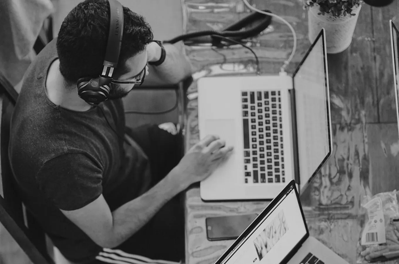 Person wearing headphones working on a laptop at a wooden table with another laptop and a smartphone nearby.