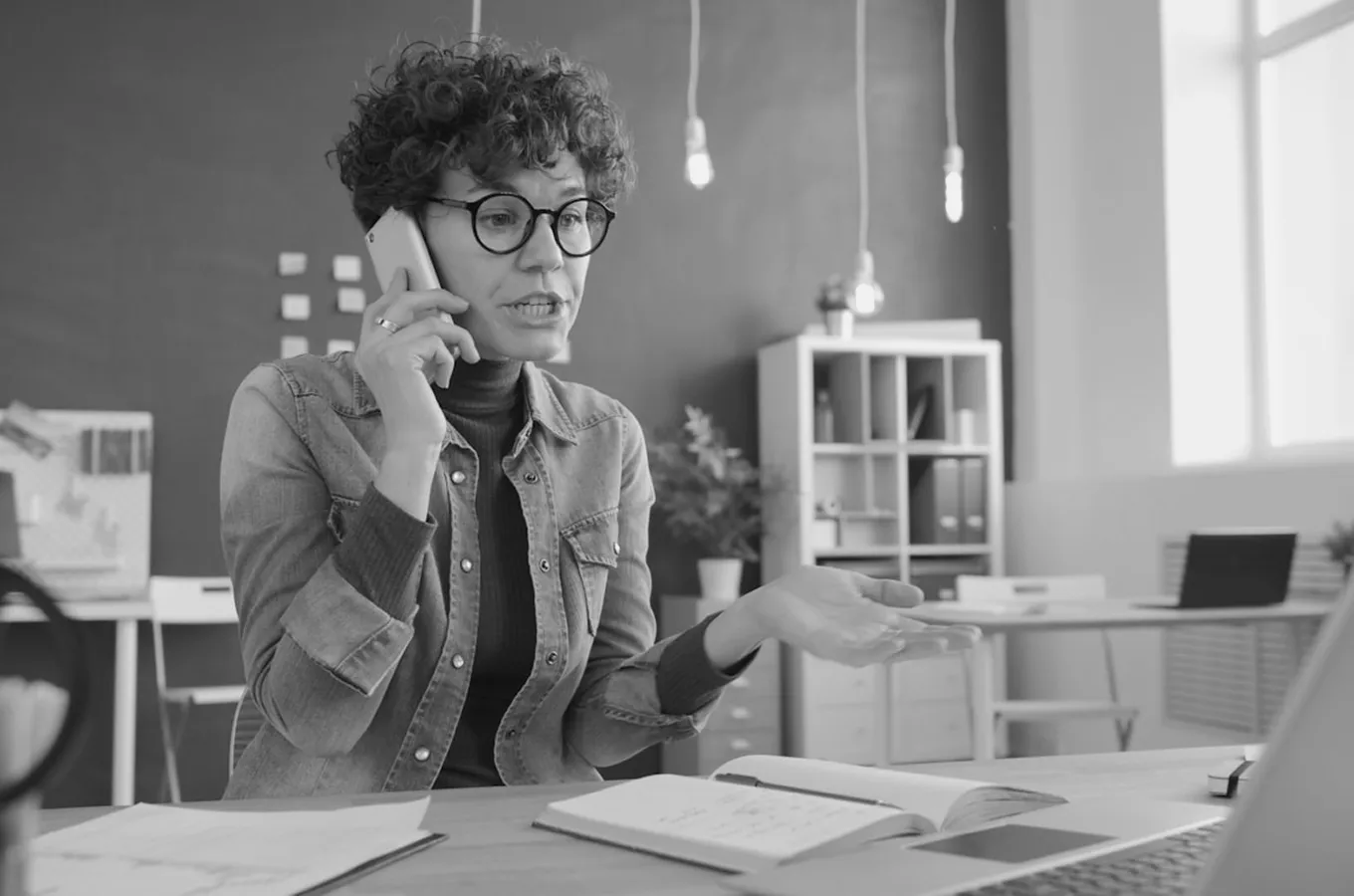 Woman with curly hair and glasses talking on a smartphone while gesturing with her hand in an office setting.