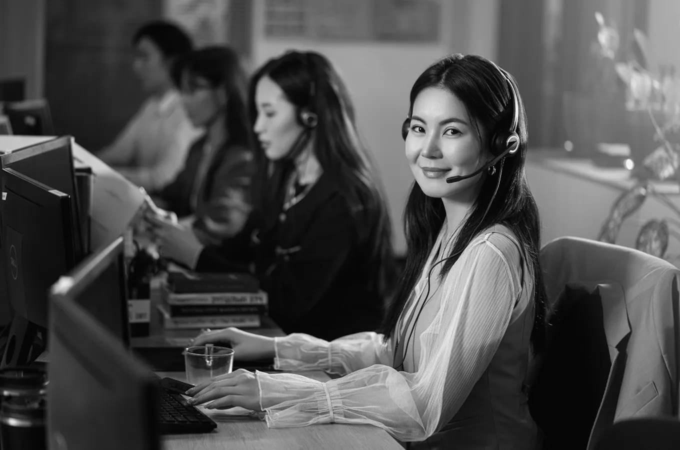 Smiling woman wearing a headset working at a computer in a busy call center with colleagues in the background.