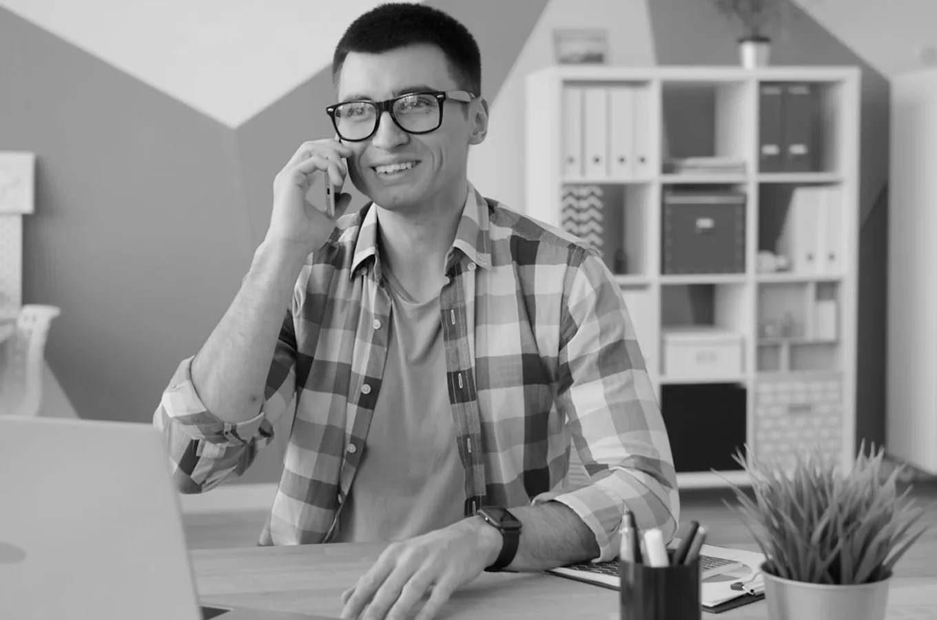 Smiling man wearing glasses and plaid shirt talking on a smartphone at a desk with a laptop, pens, and a plant.