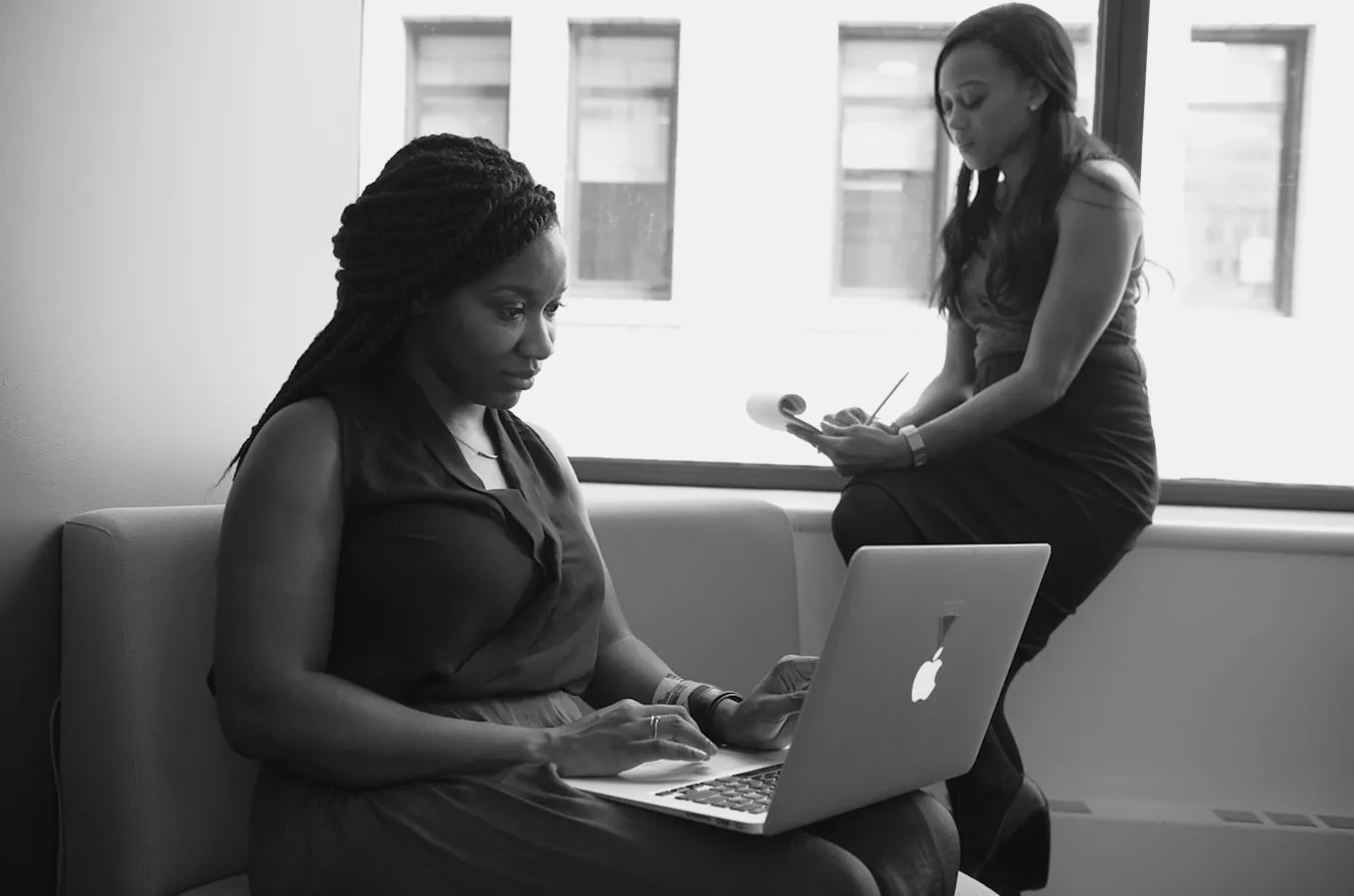 Two women working in an office, one typing on a laptop and the other writing on a notepad by a window.