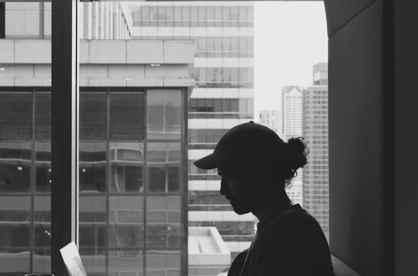 Silhouette of a person wearing a cap and working on a laptop by a large window with city buildings in the background.