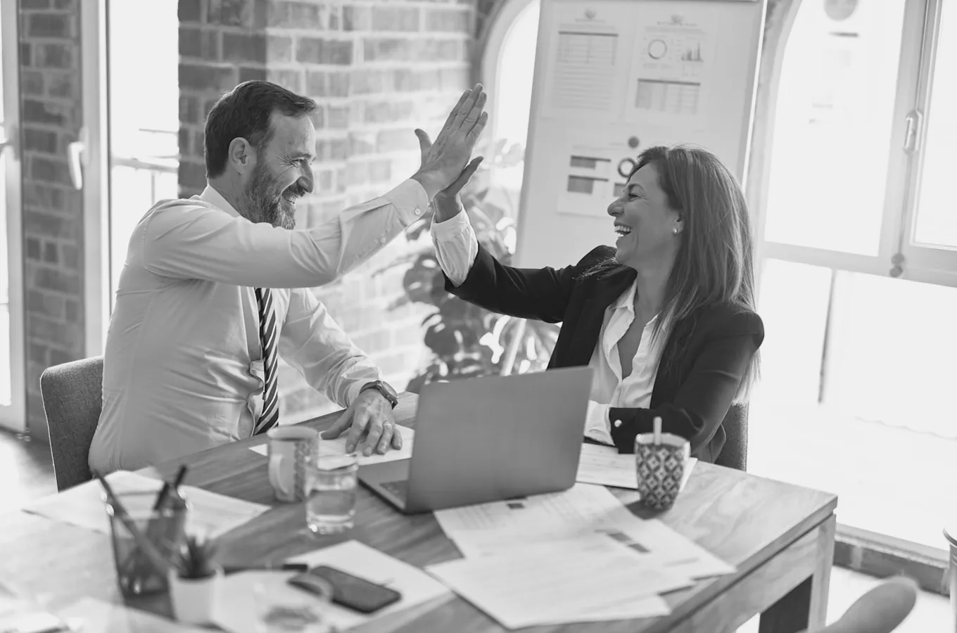 Smiling man and woman giving a high-five across a desk with a laptop and papers in an office setting.