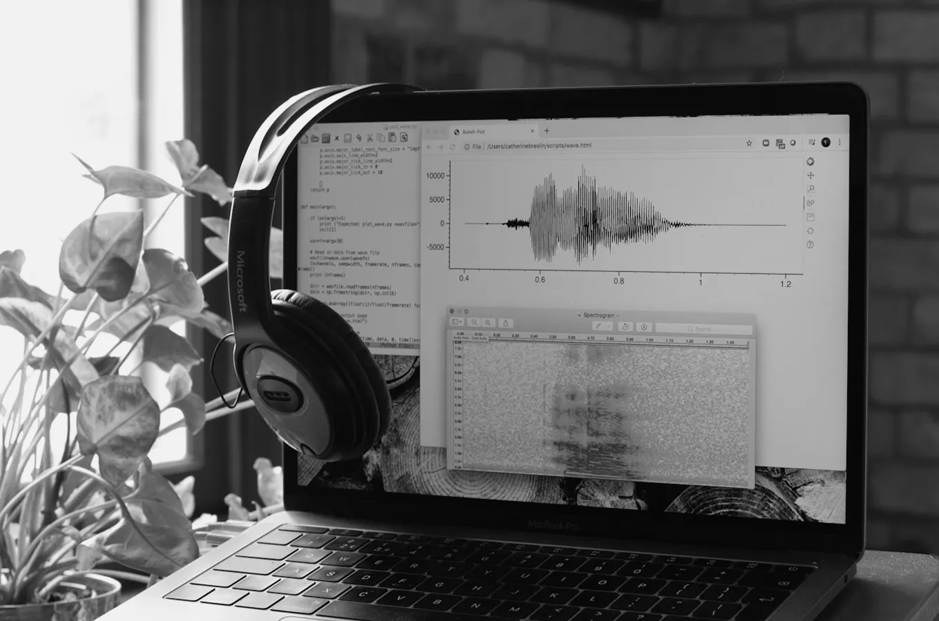 Laptop showing audio waveform and spectrogram analysis with headphones hanging on the side and a potted plant nearby.
