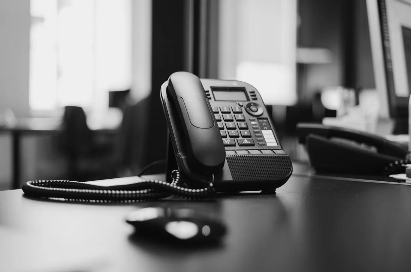 Black office desk phone with cord placed on a desk in a blurred office setting.