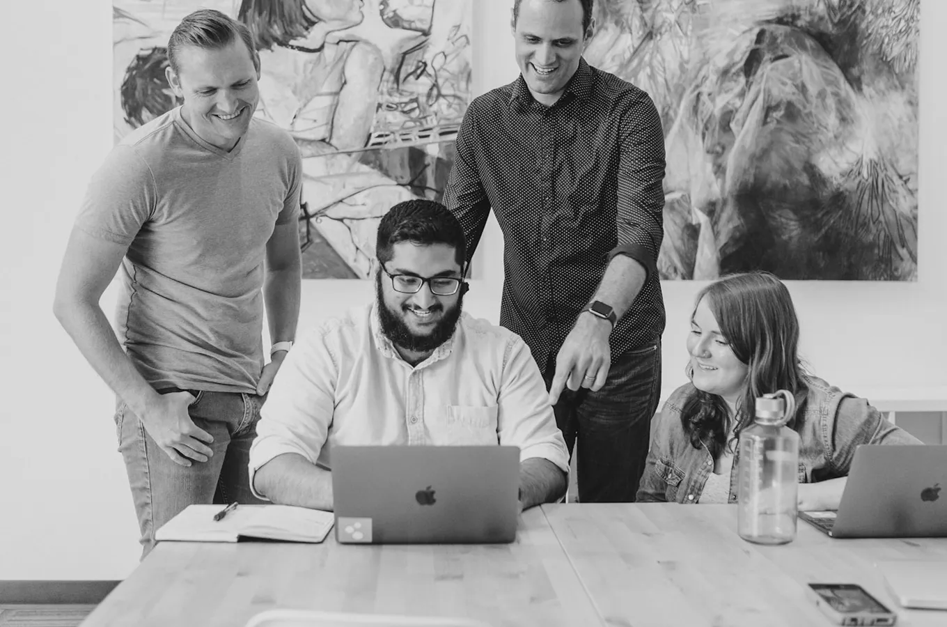 Four people collaborating around a table, smiling and looking at a laptop screen.