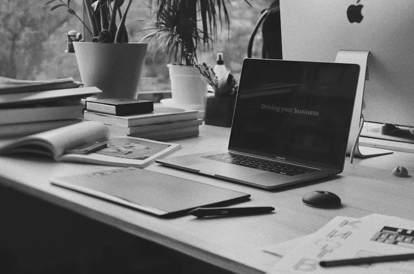 Modern workspace with a MacBook Pro laptop displaying 'Driving your business,' surrounded by books, plants, and papers on a light wooden desk.
