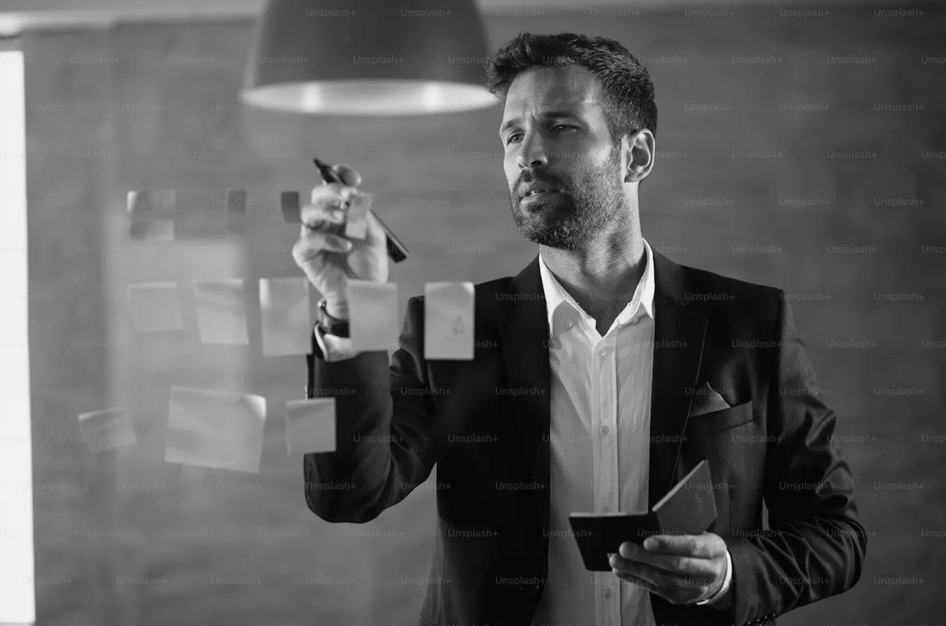 Man in suit writing on sticky notes on a glass wall while holding a notebook.