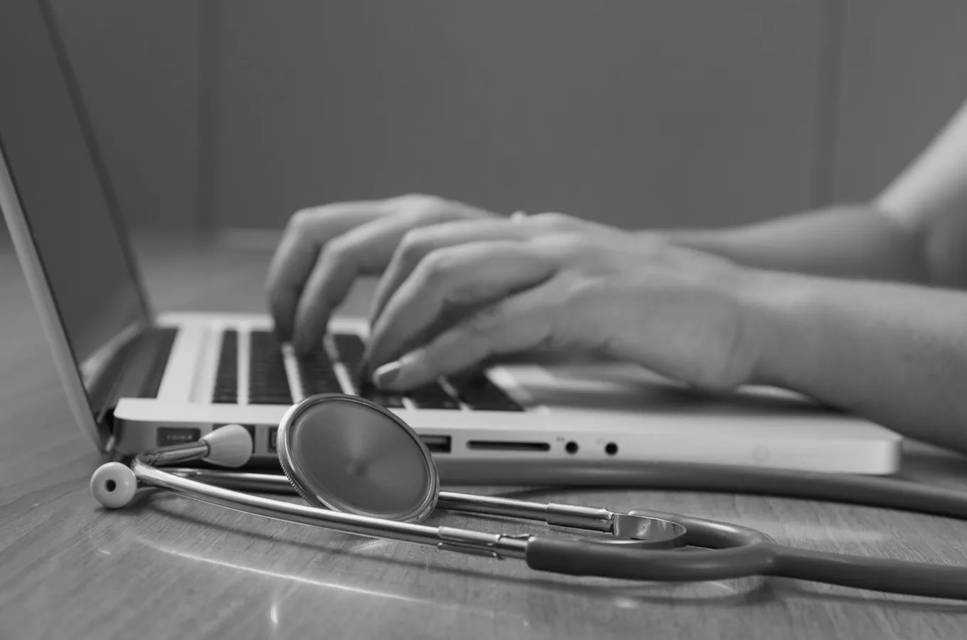 Hands typing on a laptop keyboard with a stethoscope lying on the table in the foreground.