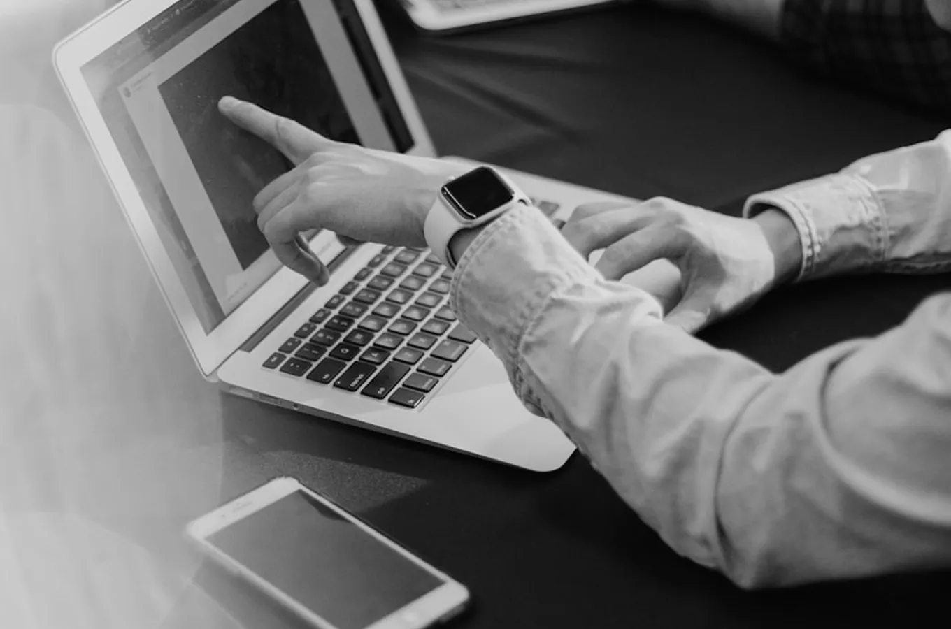 Person wearing a smartwatch pointing at a laptop screen with another hand on the keyboard and a smartphone on the table.