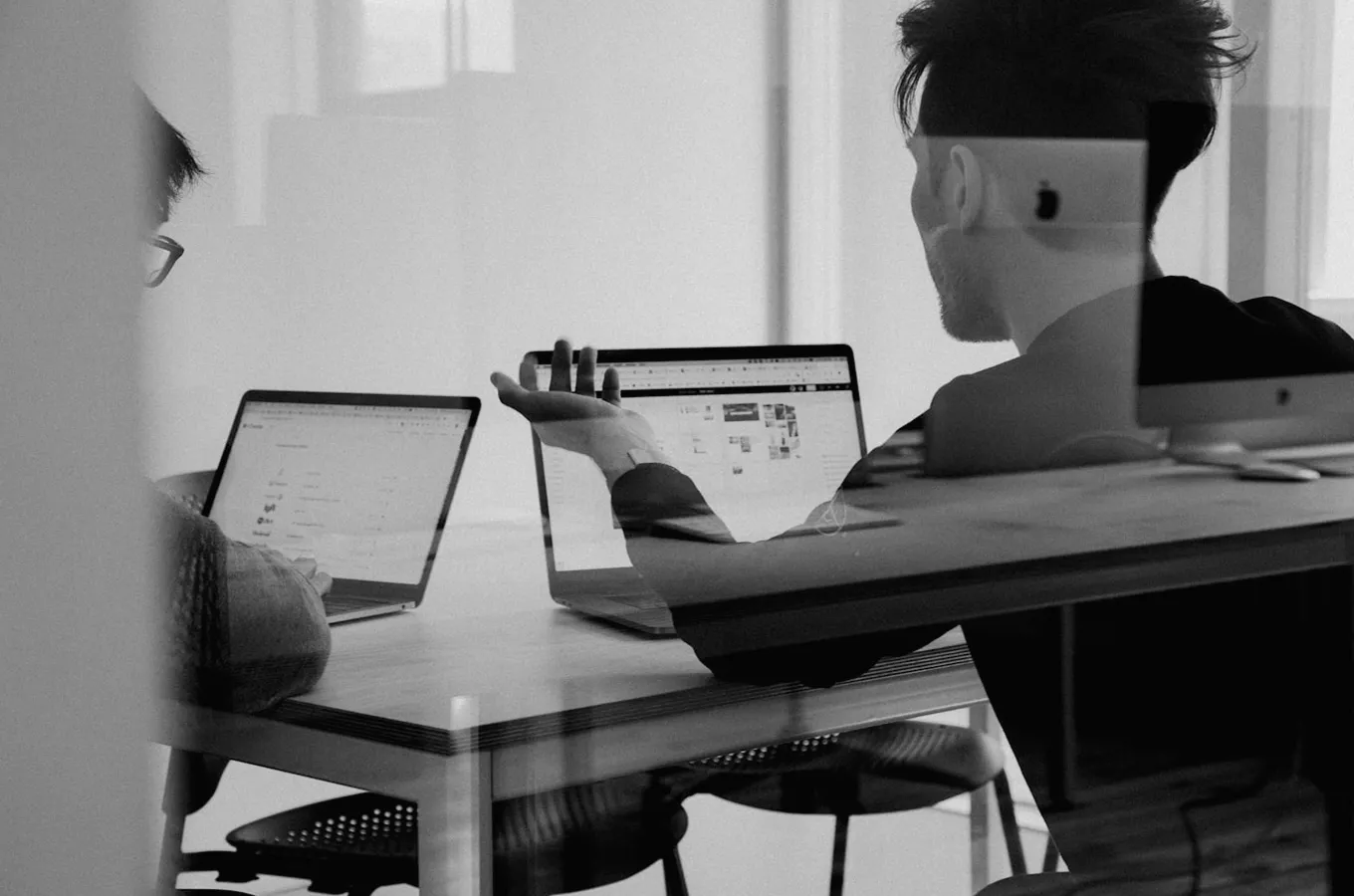 Two people sitting at a table using laptops, one person gesturing with a raised hand during discussion.