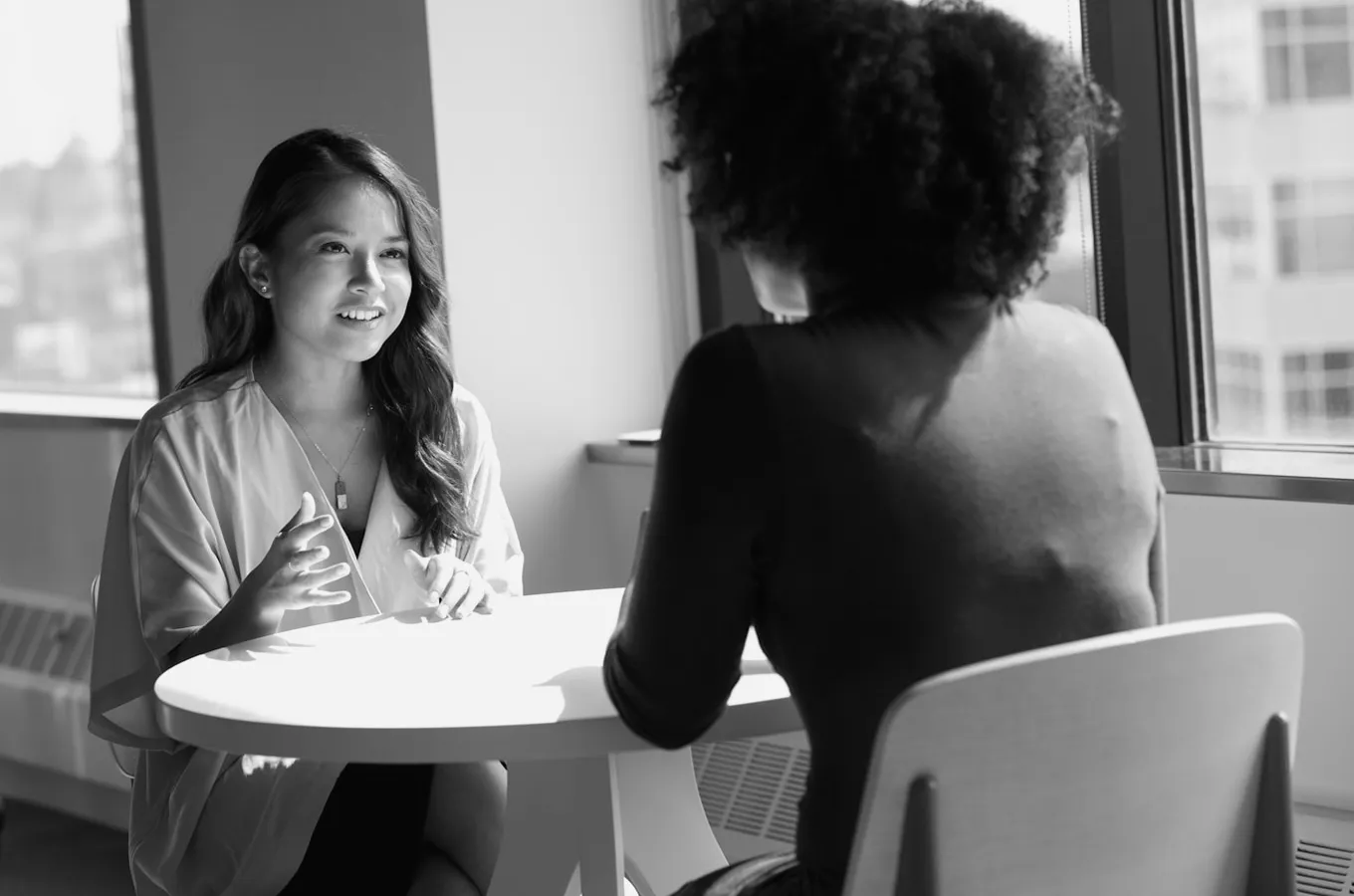 Two women having a conversation at a round table near a window, one woman facing the camera and speaking with hand gestures.