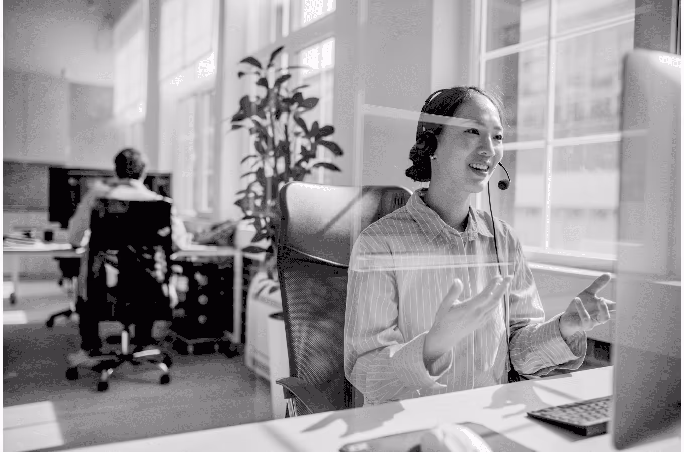 Smiling woman wearing headset and gesturing while talking in a bright office with large windows.