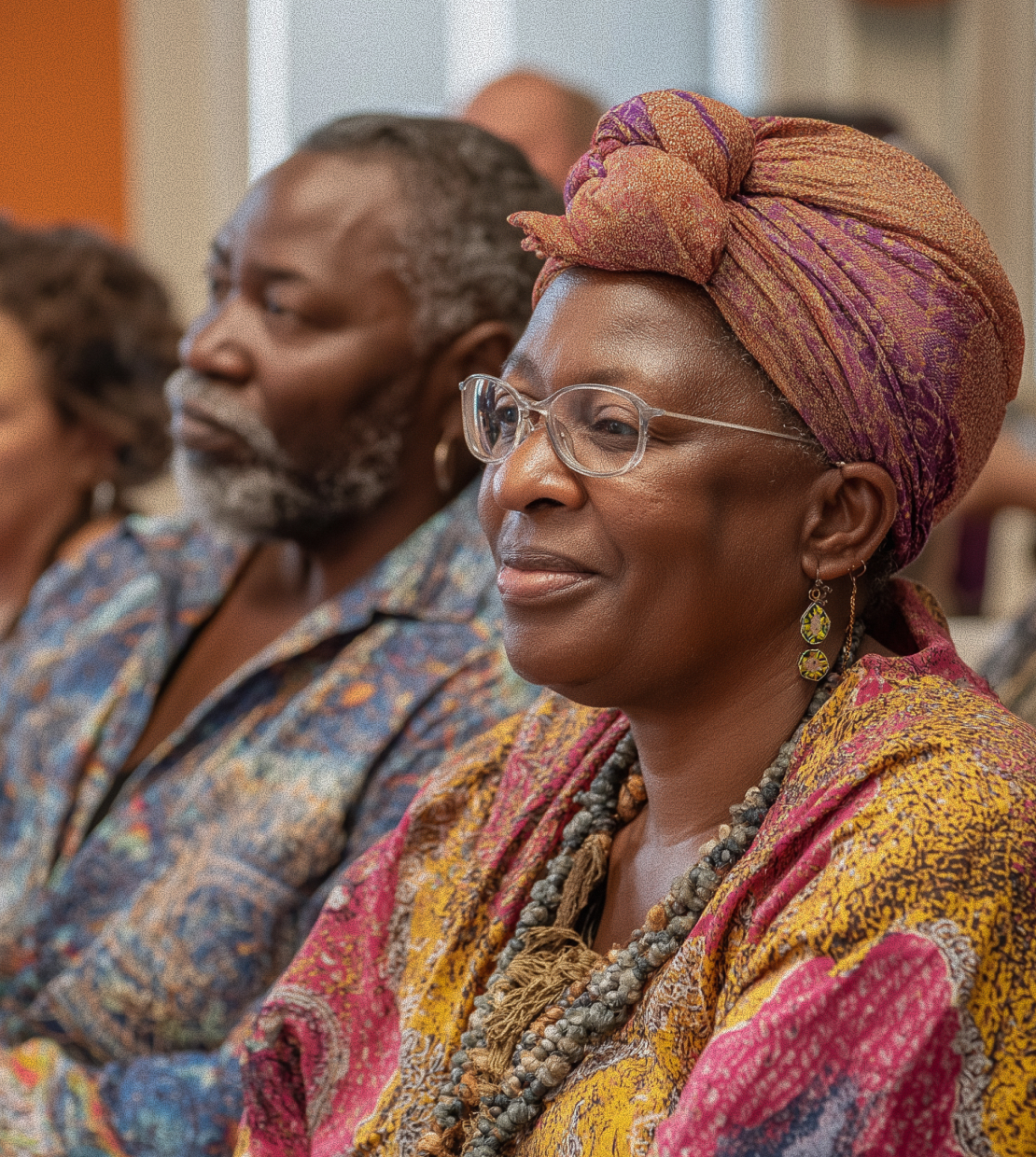 Smiling woman wearing glasses, a colorful headwrap, and patterned clothing sitting attentively in a group.