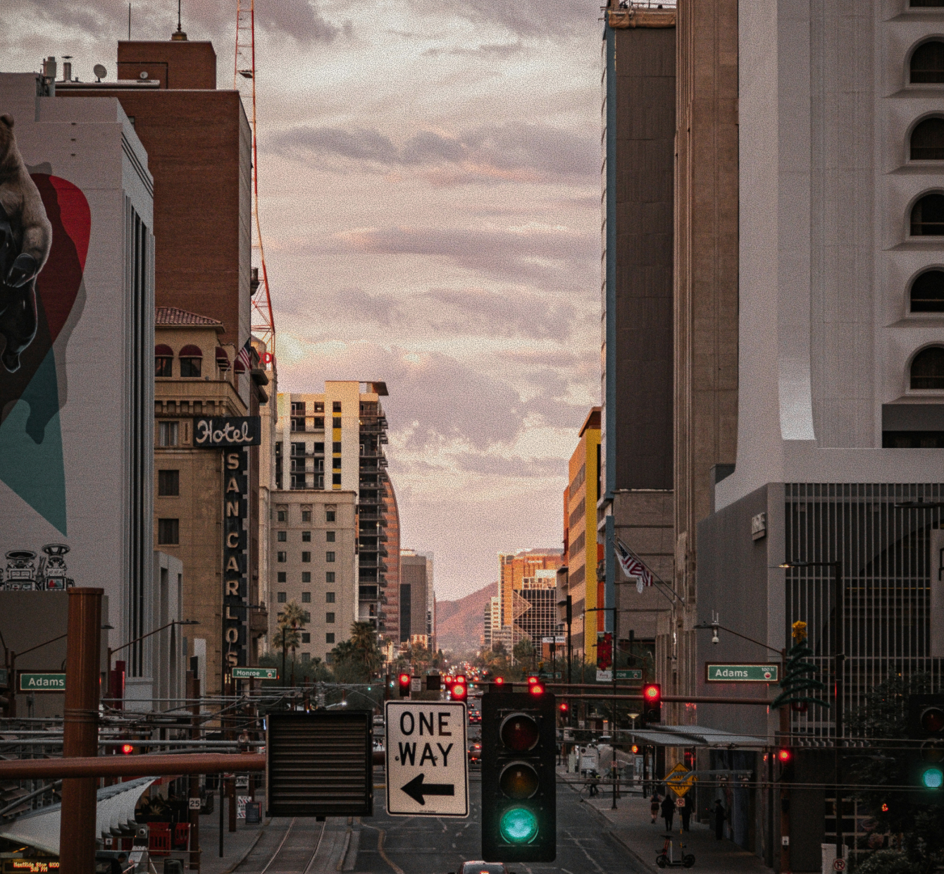 Downtown city street at dusk with traffic lights, one way sign, tall buildings, and mountains in the background.