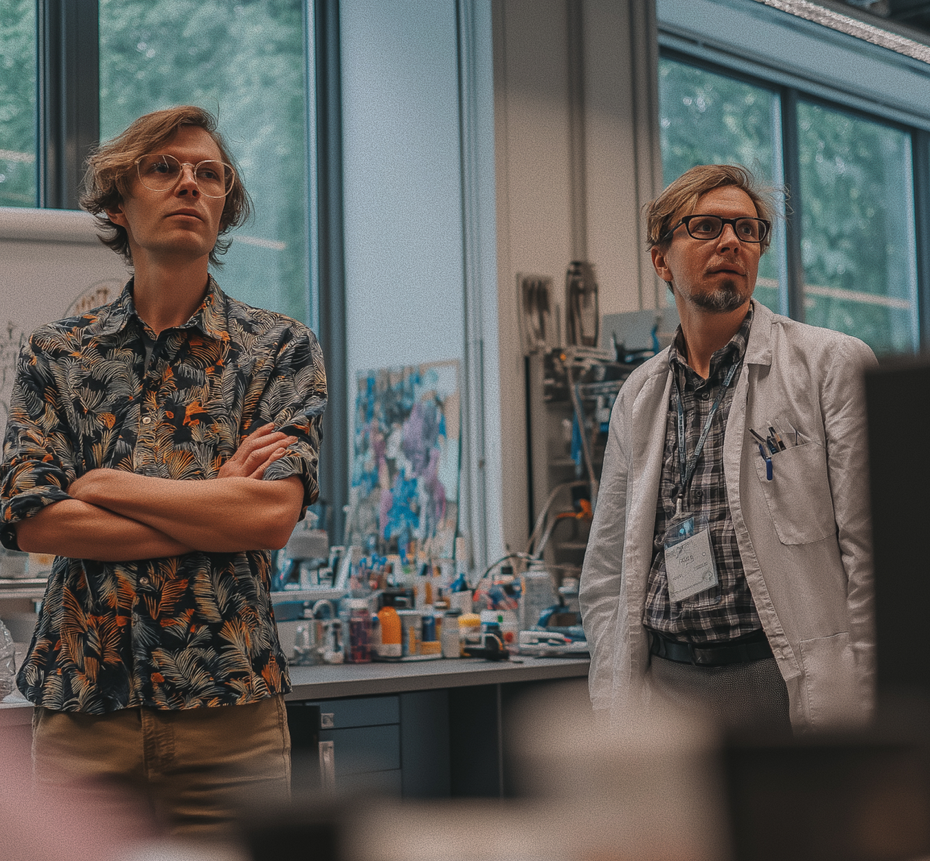 Two men in a laboratory; one in a patterned shirt with arms crossed and one in a white lab coat with pens in pocket.