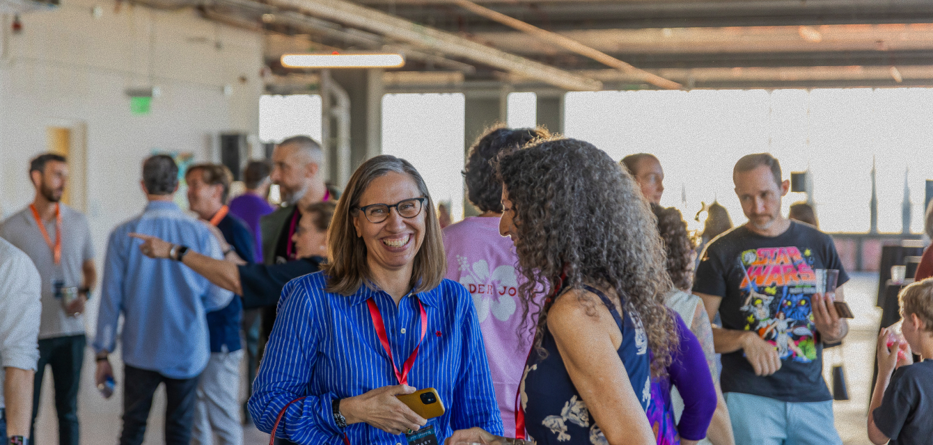 People socializing at an indoor event with two women smiling in the foreground, one holding a phone.