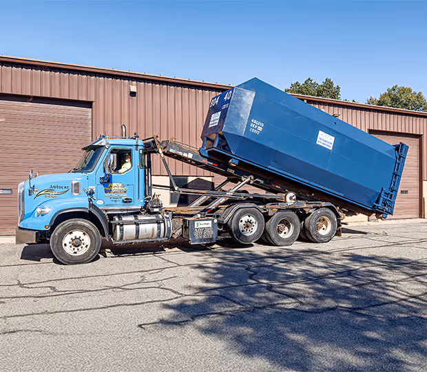 Blue rolloff truck unloading large blue container against a brown industrial building background.
