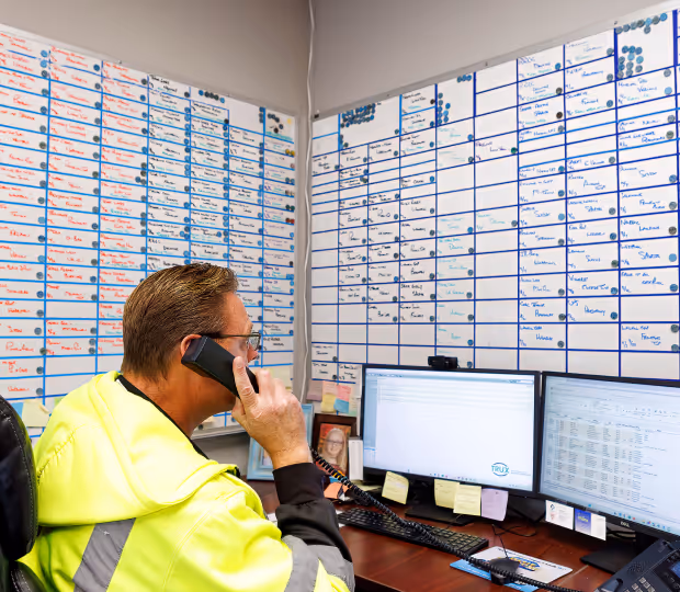Man in high-visibility jacket on phone, sitting at desk with dual monitors and scheduling boards full of notes on walls behind him.
