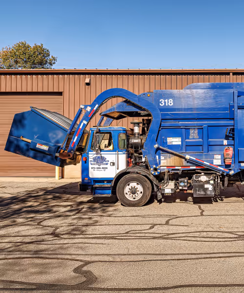 Blue waste disposal truck lifting a large blue commercial dumpster in front of a brown building under a clear sky.