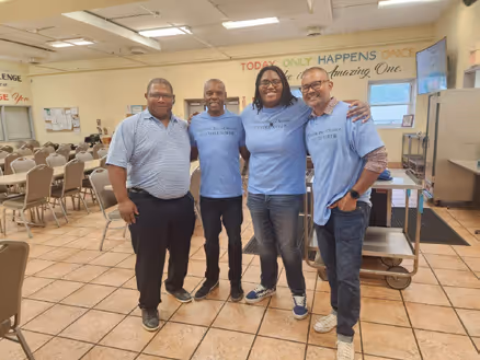 Four smiling men standing indoors in a meeting room with chairs and tables, three wearing matching light blue t-shirts.