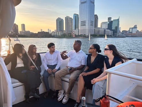 Five diverse adults sitting and talking on a boat with a city skyline and sailboats on the water at sunset.