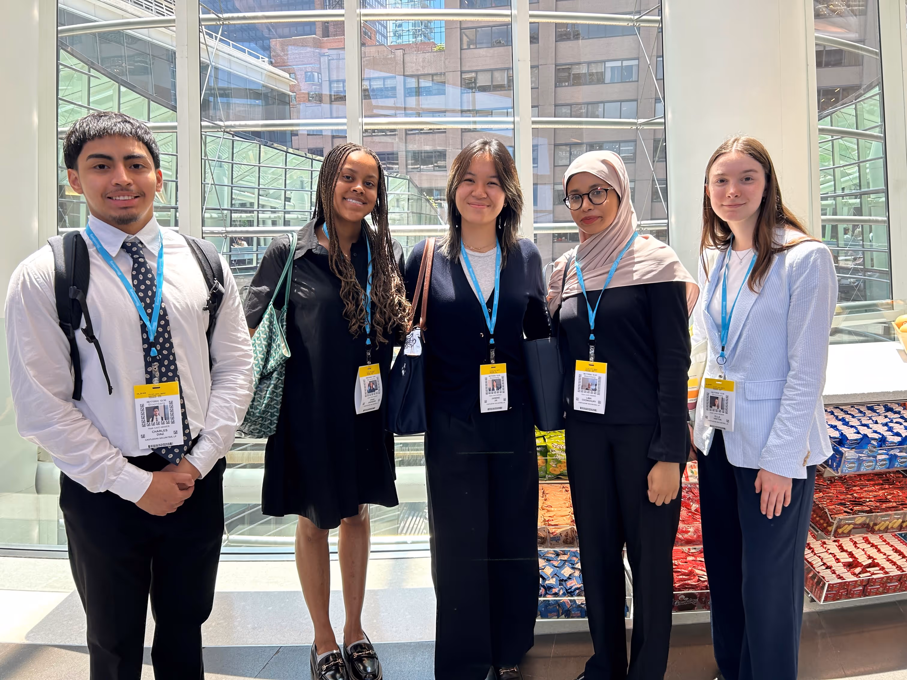 Group of five young professionals wearing conference badges and business attire standing indoors near large glass windows with city buildings visible outside.