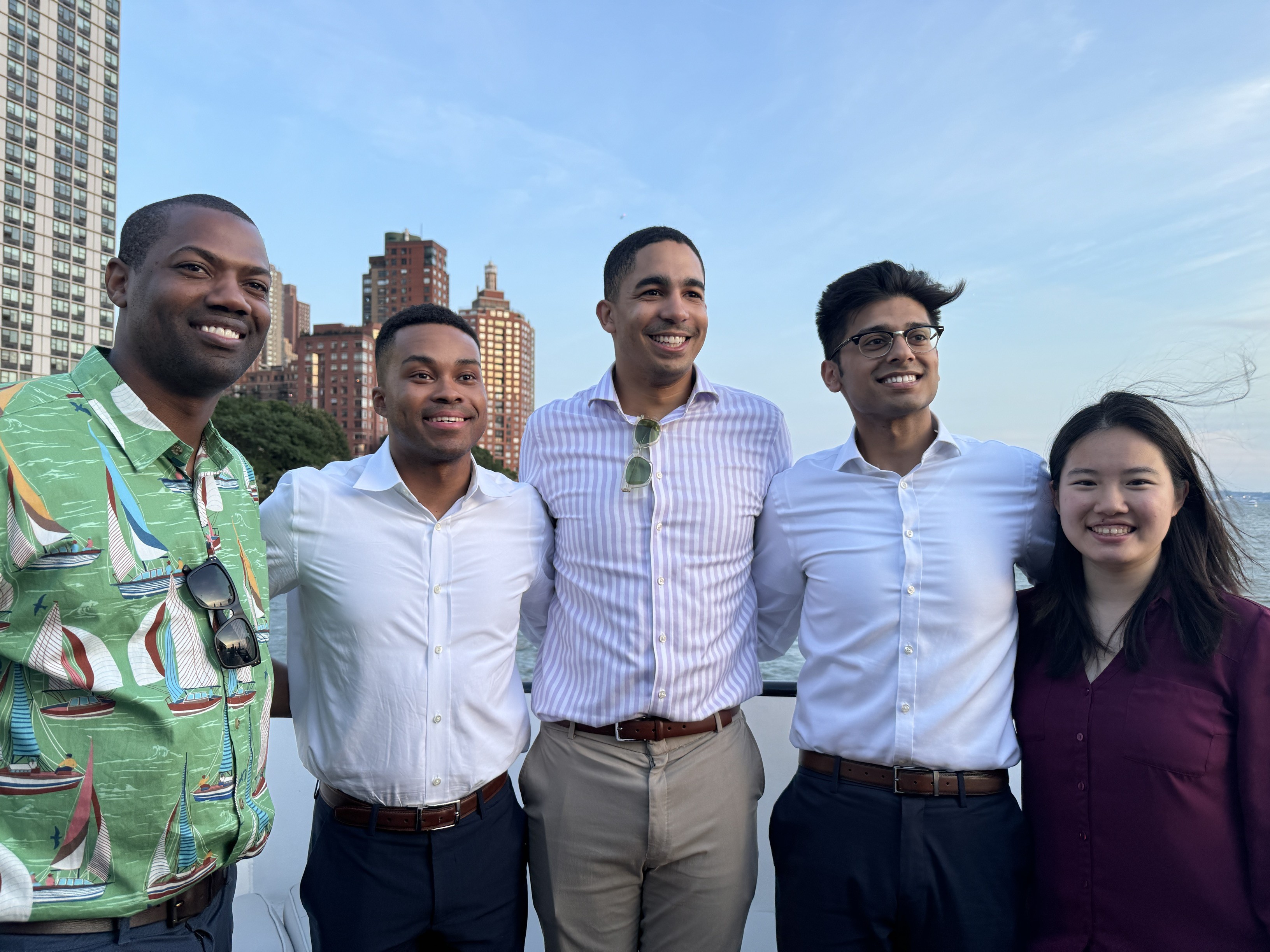 Five diverse young adults smiling and posing together outdoors in front of city buildings and water at sunset.