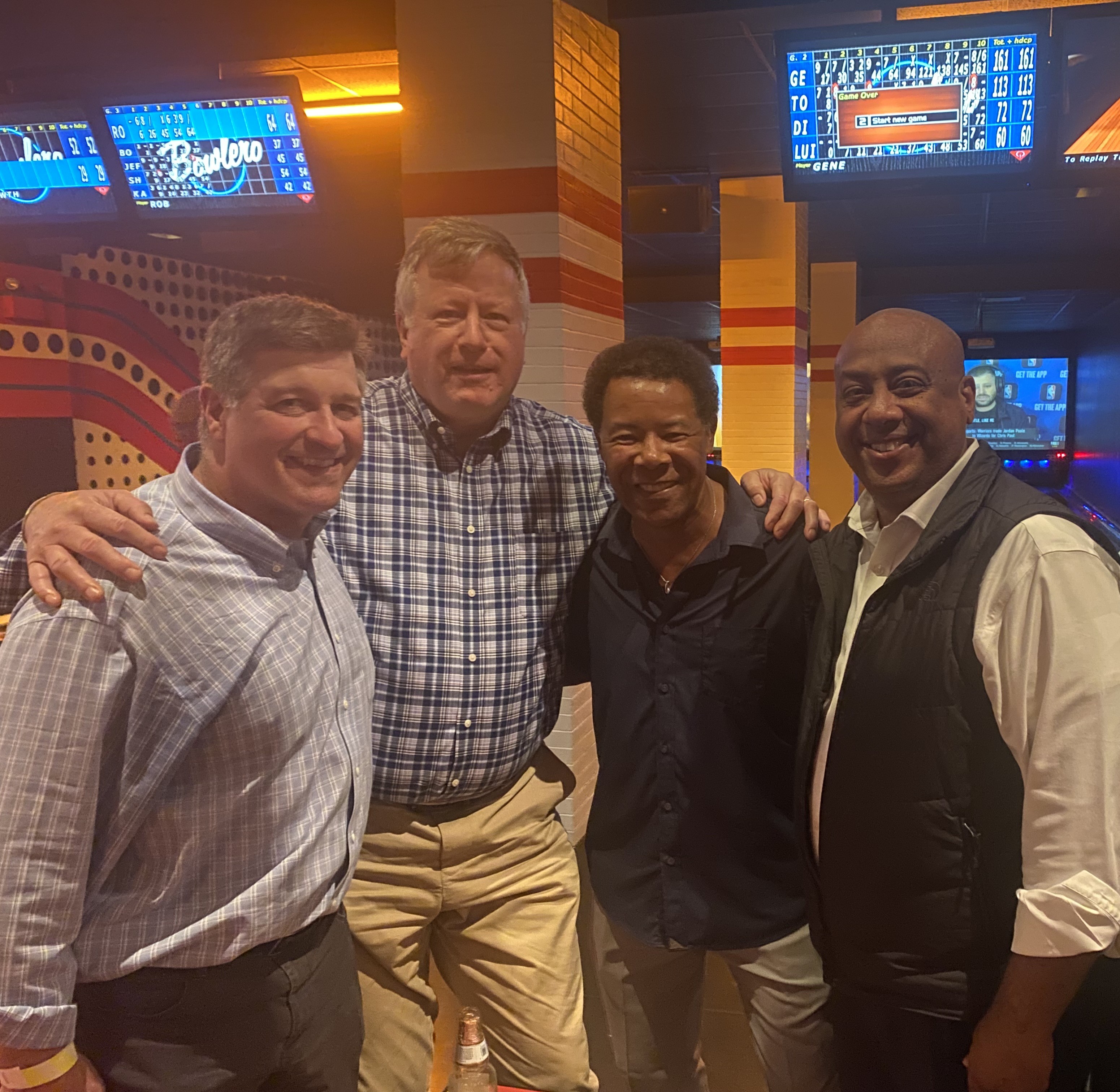Four men smiling and posing together inside a bowling alley under illuminated scoreboards.