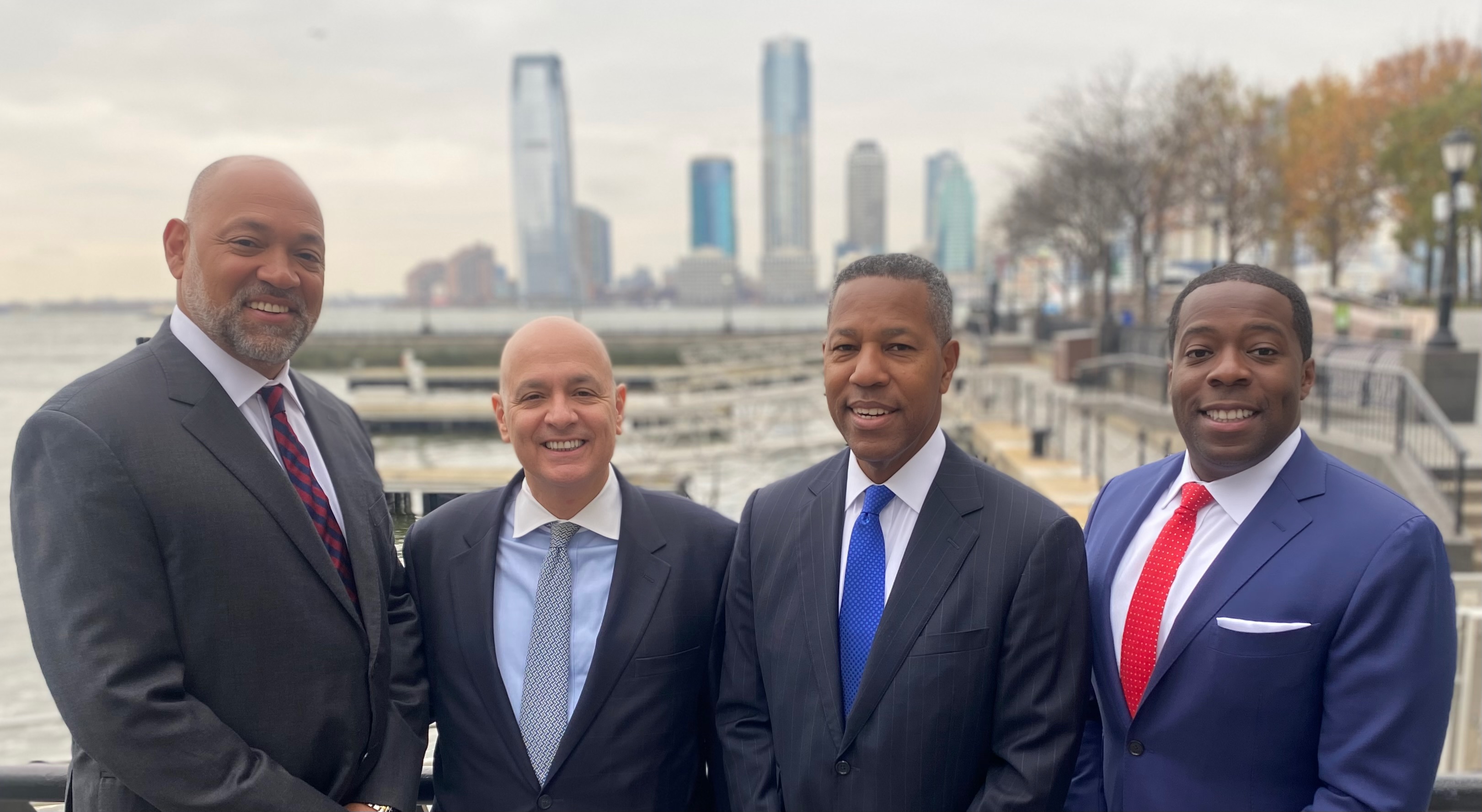 Four men in business suits smiling outdoors with a city skyline and water in the background.