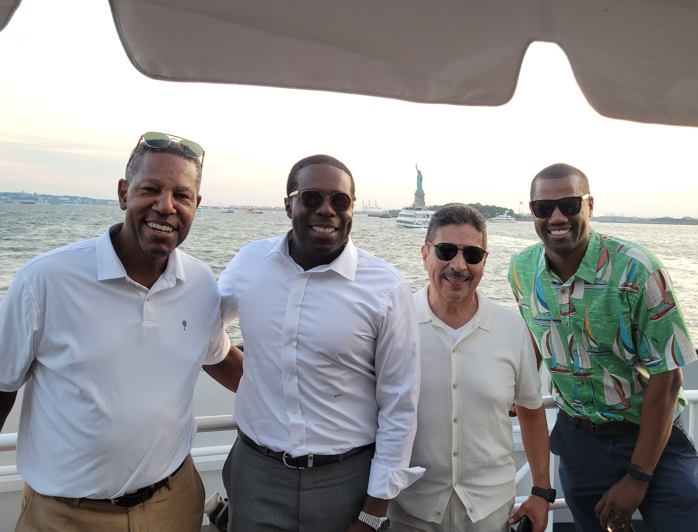 Four men smiling and posing together on a boat with the Statue of Liberty and water in the background.